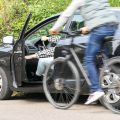 Ein Mann fährt auf einem Fahrrad an einem geparkten Wagen vorbei, dessen Fahrerin gerade ihre Tür öffnet. Die Polizei rät: Vor dem Öffnen einer Autotür immer auch den rückwärtigen Verkehr beachten. Dabei hilft der „Holländische Griff“.