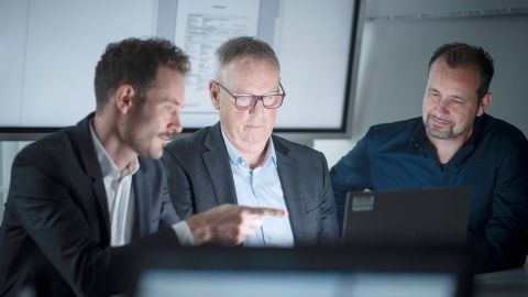 Dennis Sopp (LZPD NRW), Jürgen Marten (LKA NRW) and Michael Richter (PP Aachen) sit together in an office in front of a laptop and look at the screen. Dennis Sopp points his finger at the screen. A white wall and a large screen can be seen in the background.